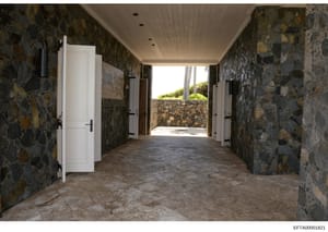 This photograph shows the interior of a covered hallway or corridor with exposed stone walls in earth tones and whites. The space features white wooden doors on both sides, recessed ceiling lights, and opens to an outdoor area with visible stonework 