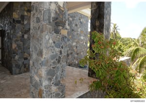 This photograph shows a covered outdoor structure featuring rustic stone columns and tropical landscaping. The image appears to be from a residential property in a tropical or subtropical location, showing architectural details and grounds that may b