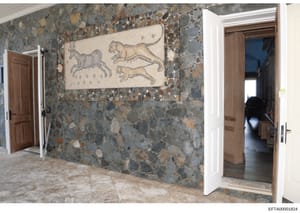 This photograph documents an interior hallway or corridor featuring a decorative mosaic tile wall and classical artwork. The space displays a sophisticated multi-colored stone or terrazzo wall with embedded tiles in earth tones, along with a framed m