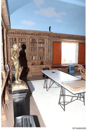 This photograph documents the interior of a residential room featuring custom wooden built-in bookshelves, large windows with orange-red treatments, and valuable art collection display. The room contains classical sculptural pieces displayed on pedes