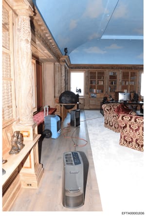 This photograph shows the interior of a luxuriously decorated room featuring elaborate architectural details, built-in wooden bookshelves, ornate furnishings, and a decorative globe. The room appears to be undergoing restoration or environmental reme