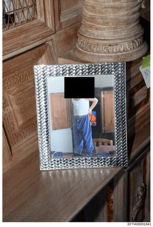 This is an evidence photograph showing a framed picture discovered during what appears to be an interior search of a property. The photograph itself is partially redacted (face obscured with black redaction bar). The frame is ornate with a metallic b