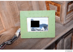 This is an evidence photograph showing a framed photograph displayed on a wooden surface. The inner photograph contains a mountain landscape scene with snow-covered peaks visible in the background, though a portion of the image is redacted with a bla