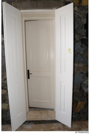 This is an evidence photograph depicting an interior doorway with white painted doors and frame. The image shows a two-door entrance opening into a room with stone or textured dark walls visible on either side. A yellow evidence marker or tag is visi