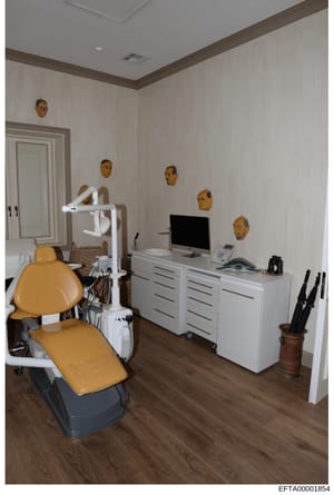 This photograph shows an interior view of what appears to be a dental office or treatment room, featuring a yellow dental chair, overhead lamp, computer workstation, and white cabinetry. The room is decorated with multiple golden-colored decorative f