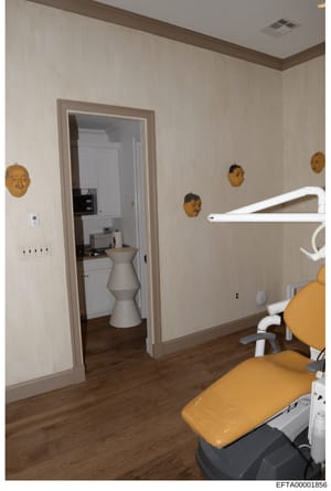 This photograph shows an interior view of what appears to be a dental or medical office space. The room features modern furnishings including a dental chair with yellow upholstery in the foreground, white equipment, decorative gold-colored masks moun