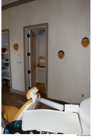 This photograph documents an interior room featuring white walls with decorative wooden mask sculptures mounted on the walls. The room contains a doorway with dark framing, electrical outlets, and appears to be part of a larger residence. The image w