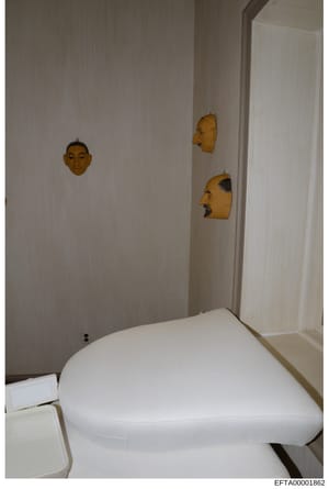 This is an evidence photograph from an Epstein property showing a bathroom interior. The room features three decorative wooden masks hanging on light-colored walls above a white toilet. The masks appear to be carved wooden art pieces in varying style