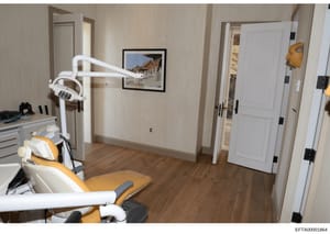 This photograph documents a dental treatment room featuring modern clinical equipment, including a dental chair with yellow upholstery, overhead surgical lighting, and cabinetry. The room has cream-colored walls, white trim, wooden flooring, and fram