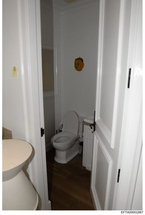 This is a photograph of a bathroom interior showing a toilet, sink, and white-painted walls with dark flooring. The image appears to be evidence documentation from a property inspection or seizure, with evidence markers visible on the walls. The Bate
