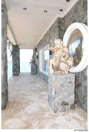 This is an interior photograph of a luxurious coastal property showing a long hallway with stone walls, high white ceiling with recessed lighting, and decorative classical sculptures. The hallway opens to ocean views, featuring ornamental urns and cl