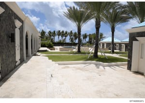 This photograph shows the exterior courtyard area of what appears to be a luxury residential property, featuring a paved terrace, manicured lawn, palm trees, a swimming pool, and white stone outbuildings with arched openings. The image documents the 