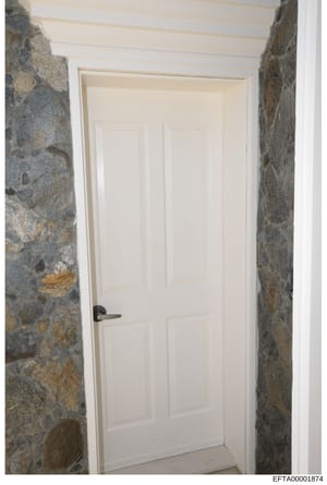 This photograph shows an interior passageway or corridor featuring rustic stone walls in gray and tan tones with a white six-panel door at the far end. The image appears to be documentation of interior architectural features or property conditions, l