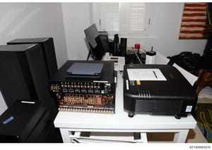 This photograph documents a residential workspace or media room containing professional-grade audio and video equipment. The setup includes an audio mixer/amplifier with multiple channels, a projector, computer monitors, laptops, and various cables a