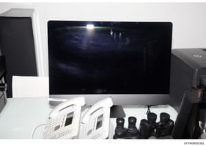 This photograph shows a desktop computer setup labeled 'KITCHEN MAC' with what appears to be surveillance or monitoring equipment, including two white IP phones/devices and multiple camera lenses. The image documents the physical equipment configurat