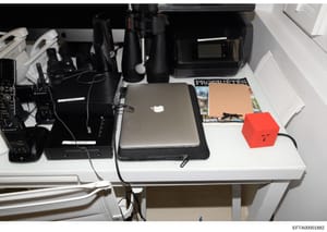 This is a photograph of a white desk containing various electronic equipment, surveillance devices, a laptop computer, and printed materials including what appears to be a magazine or publication labeled 'PROPHETES'. The image documents the physical 