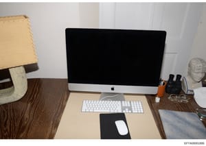 This is an evidence photograph showing a residential or office desk setup featuring an Apple iMac computer with peripherals. The image documents the physical environment and equipment, likely seized or photographed during the investigation. The desk 