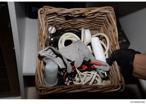 This is an evidence photograph showing the contents of a wicker basket seized during the investigation. The basket contains various household items including cleaning supplies, cords, cables, gloves, and other miscellaneous objects. The photograph wa