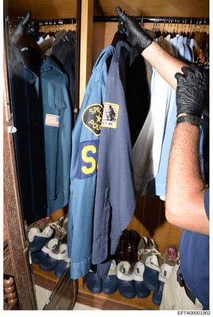This photograph shows a law enforcement officer wearing protective gloves examining a police uniform displayed in a closet during what appears to be an evidence collection or property search. The uniform bears a visible badge or patch and is hung amo