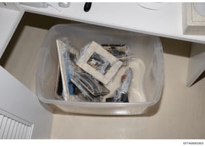 This is an evidence photograph showing a clear plastic storage container filled with various wrapped items, photographs, and documents. The contents appear to be preserved evidence, with multiple items wrapped in plastic. The photo was taken as part 