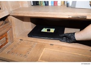 This photograph shows a law enforcement evidence photo documenting a hidden compartment discovered in a wooden desk. An investigator wearing black gloves is holding open a concealed drawer or compartment beneath the desk surface, revealing what appea