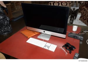 This is an evidence photograph showing a desk workspace containing an Apple iMac computer, wireless keyboard and mouse, orange notebook, sunglasses, a scented candle, audio equipment, and other personal items on a red desk surface. The photo document