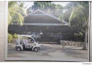 This is a photograph of a residential property featuring a large covered structure with a peaked roof, surrounded by tropical vegetation including palm trees. A white golf cart is parked in the foreground on a paved driveway. The image appears to be 