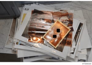 This photograph shows a collection of photographic negatives and contact sheets stacked on a marble surface, including film strips in protective sleeves and what appears to be a wooden-framed negative or slide with two redacted sections. The image do