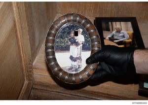 This is an evidence photograph showing a framed oval portrait being held by an investigator wearing black gloves. The framed photo depicts two young people outdoors with foliage in the background, and one face is redacted with a black block. In the b
