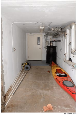 This is an interior photograph of a basement or utility room showing storage and mechanical equipment. The space contains a red-orange kayak, a yellow hand truck/dolly, various plumbing and HVAC pipes, a black storage tank, shelving with equipment, a
