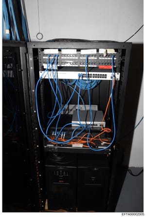 This photograph shows a black server rack containing networking equipment, switches, and cabling infrastructure. The rack features blue and orange network cables organized throughout multiple tiers of equipment, with an APC power management unit visi