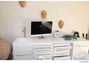 This photograph shows a contemporary bedroom or office space featuring a white desk with integrated sink, an Apple iMac computer, office equipment including a telephone and binoculars, and distinctive decorative elements including three gold-colored 