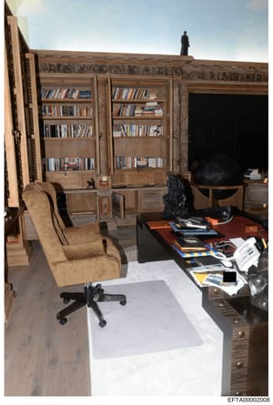 This is a photograph of an office or study room featuring built-in wooden bookshelves, antique furniture, and a desk with scattered items. The room displays ornate wood paneling and architectural details consistent with a high-end residence or proper
