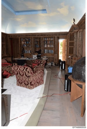 This photograph depicts a luxurious interior room featuring floor-to-ceiling wooden bookshelves, ornate wood paneling, and elaborate furnishings including burgundy patterned upholstered furniture. The room appears to be under construction or renovati