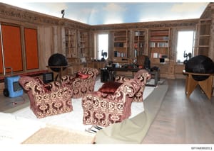 This photograph shows an elegant library or study room featuring floor-to-ceiling wooden built-in bookshelves, ornate patterned armchairs upholstered in burgundy and cream fabric, black decorative globes, and multiple windows. The space appears to be