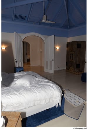 This is an evidence photograph depicting an interior bedroom space with distinctive blue ceiling and white walls. The room contains a bed with white bedding, an arched doorway, wall sconces, and a ceiling fan. The photograph appears to be part of the