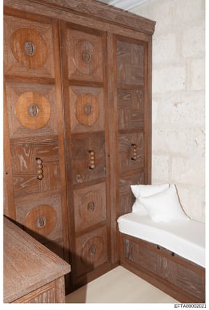 This is a photograph of an interior bedroom space, showing a large wooden wardrobe or armoire with decorative circular inlays and brass handles, positioned next to a bed with white linens. The image appears to be from a property documentation or evid