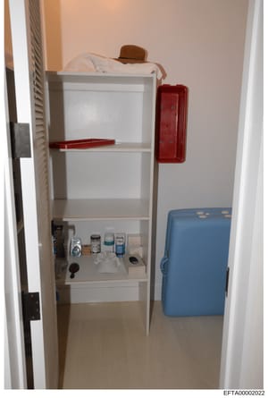 This is an evidence photograph depicting the interior of a residential closet containing white shelving units with various personal items and toiletries on the lower shelf, a red emergency phone box on the wall, a brown hat and bedding on the upper s