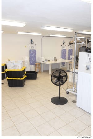 This is a photograph of a large institutional or commercial laundry/utility room, likely from an Epstein property. The space contains multiple ironing boards, storage containers, cleaning supplies, and household equipment including a standing fan and