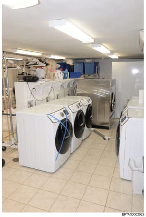 This photograph depicts a modern commercial or institutional laundry room, featuring multiple front-loading washing machines and dryers arranged in rows. The facility is equipped with industrial-grade appliances, overhead lighting, and storage shelvi