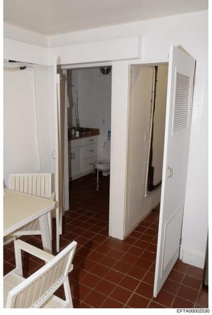 This photograph shows the interior of a kitchen and connected bathroom space, featuring white cabinetry, dark tile flooring, and various household appliances. The image appears to be evidence documentation from a property inspection or seizure relate