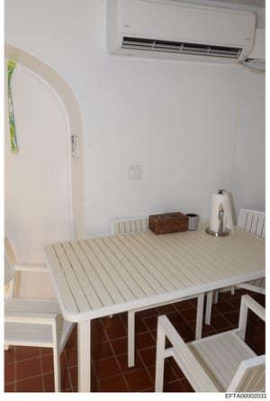 This is a photograph of a small residential room or living space, featuring minimal furnishings including a white table with striped surface, white chairs, and basic household items. The room has white walls, a wall-mounted air conditioning unit, and