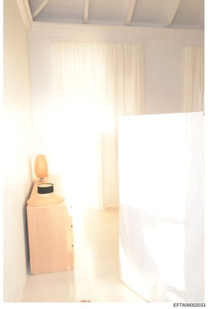 This is a photograph of a bright, minimalist interior space featuring white walls, cream-colored curtains, and minimal furnishings. A single woven table lamp with a dark band is visible against the left wall. The room appears to be a bedroom or dress
