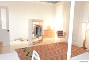 This is a photograph of an interior bedroom space, showing modern furnishings including a wooden closet organizer with hanging clothes, a wooden dresser, an upholstered chair, and a geometric patterned area rug in red and tan tones. The room features