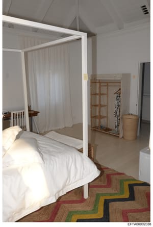 This is a photograph of a bedroom interior featuring a white four-poster bed with sheer curtains, wooden clothing rack, and minimal furnishings. The room has high ceilings, neutral walls, and a geometric patterned area rug. The purpose and context of