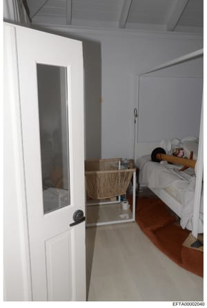 This is an evidence photograph showing an interior bedroom space with an en-suite bathroom visible through an open door. The room contains a bed with white linens, wooden headboard, and various personal items, along with a woven chair and furnishings