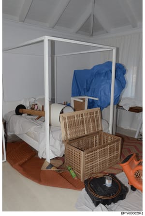 This is an evidence photograph showing a bedroom interior with a white four-poster bed, wooden storage chest, various household items, and a blue tarp or covering. The room features a vaulted ceiling, patterned area rug, and appears to be part of a r