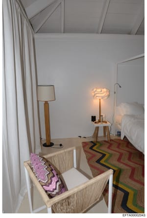 This is a photograph of a minimalist bedroom interior showing a neatly furnished room with contemporary design elements. The space includes a bed with white linens, woven furniture pieces, geometric patterned rug and pillow, wooden floor lamps, and c