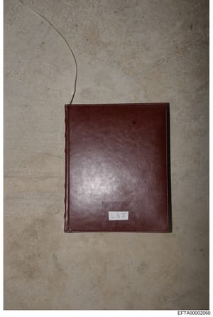 This is an evidence photograph showing a dark brown leather-bound item, possibly a journal, notebook, or similar object, photographed flat against a light textured surface. A white label bearing the initials 'LSJ' is visible on the lower portion of t