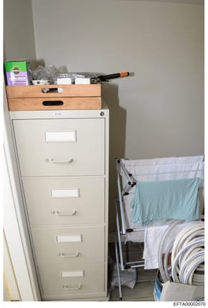This is an evidence photograph showing a corner of a room containing a white four-drawer filing cabinet (brand: Uline), wooden storage drawers on top, cleaning supplies, cooking utensils, and a laundry drying rack with linens. The image documents the